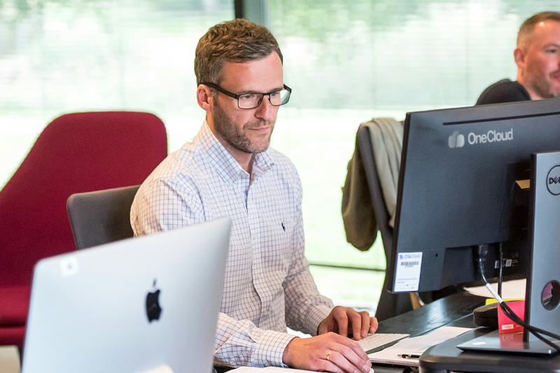 A man wearing glasses and a checkered shirt is sitting at a desk and working on a computer. Another person is visible in the background. The office is well-lit with natural light coming through large windows.